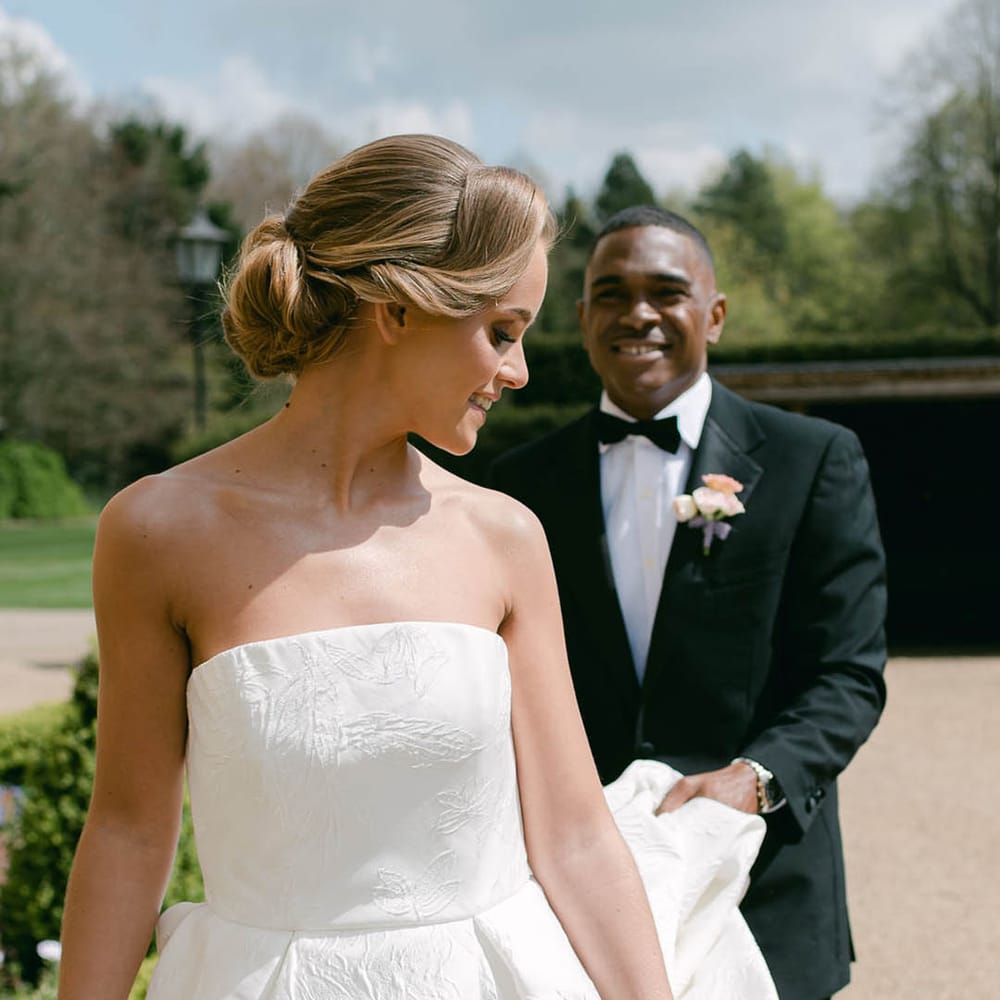 Bride with an intricate updo looks back at the groom as he lifts her wedding dress train as they walk 