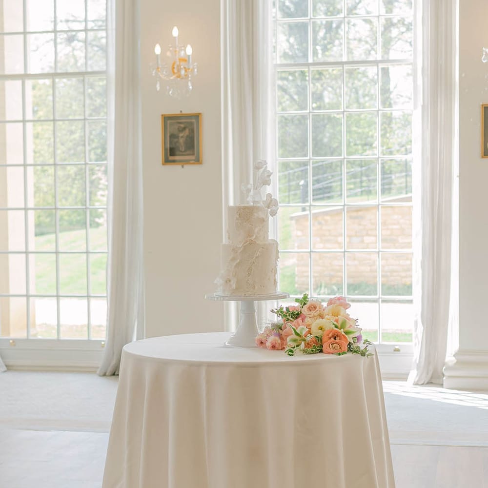 White two tier wedding cake with texture detail on a white table with the pretty pink and purple pastel bouquet 