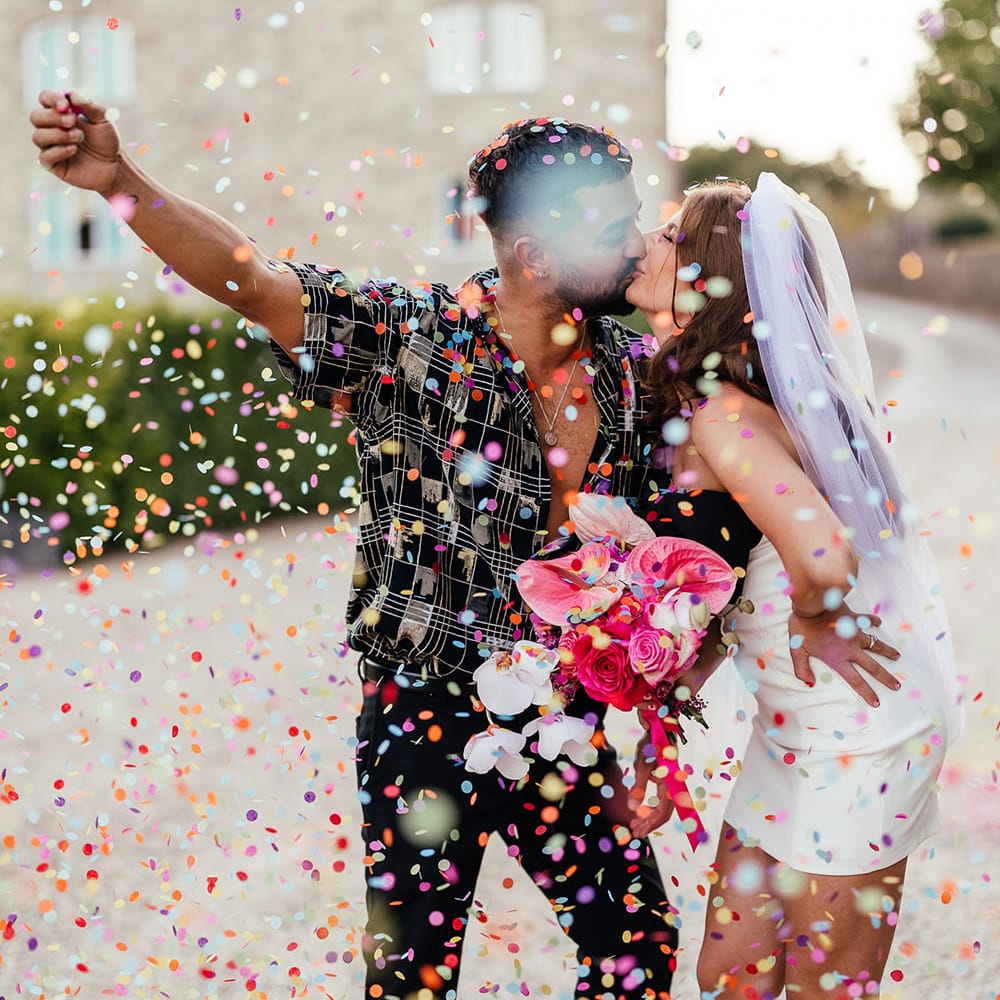 Groom pops colourful confetti around them as they kiss outdoors on their wedding day