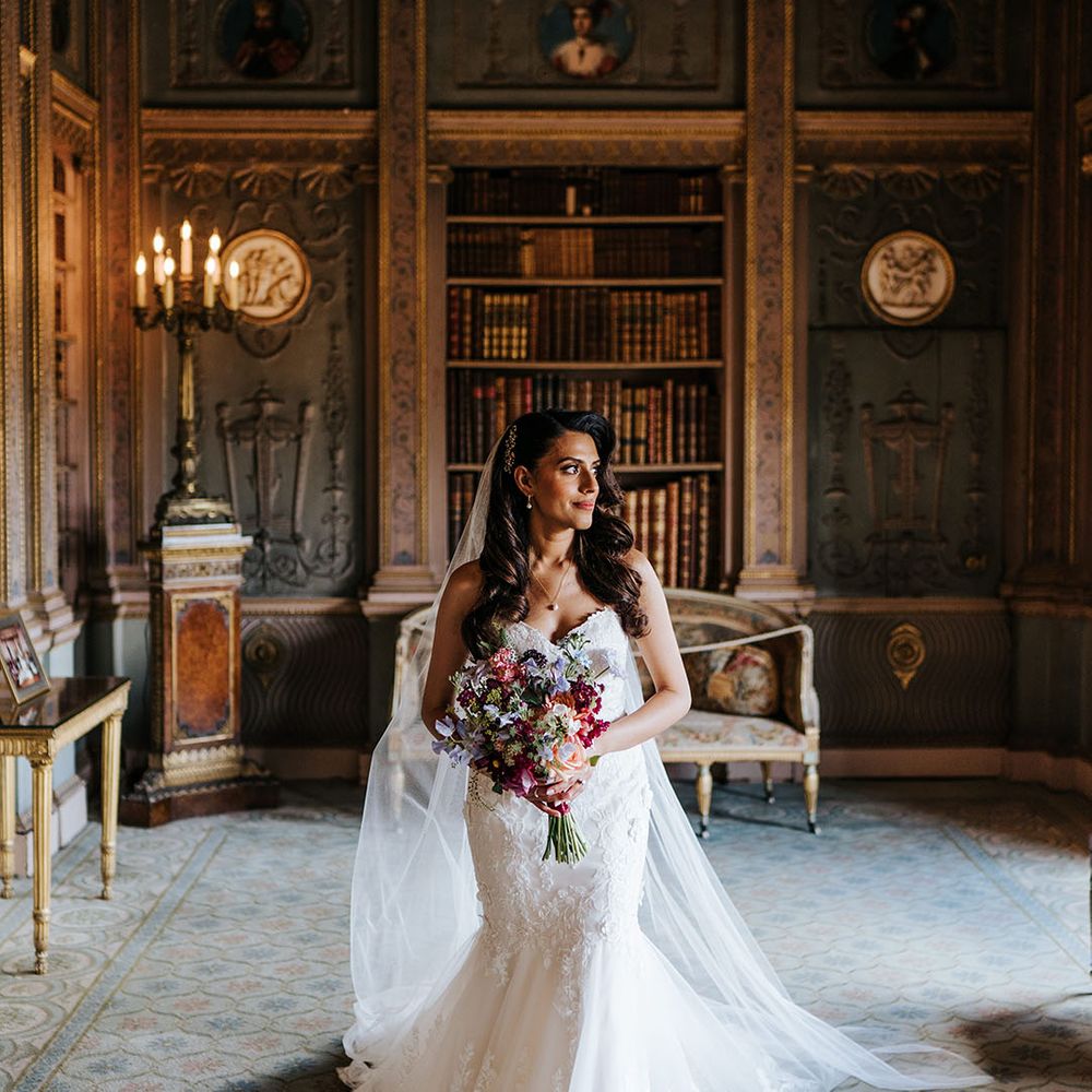 Indian bride wearing lace wedding dress with sweetheart neckline and wears her brown hair in loose waves whilst holding brightly coloured floral bouquet 