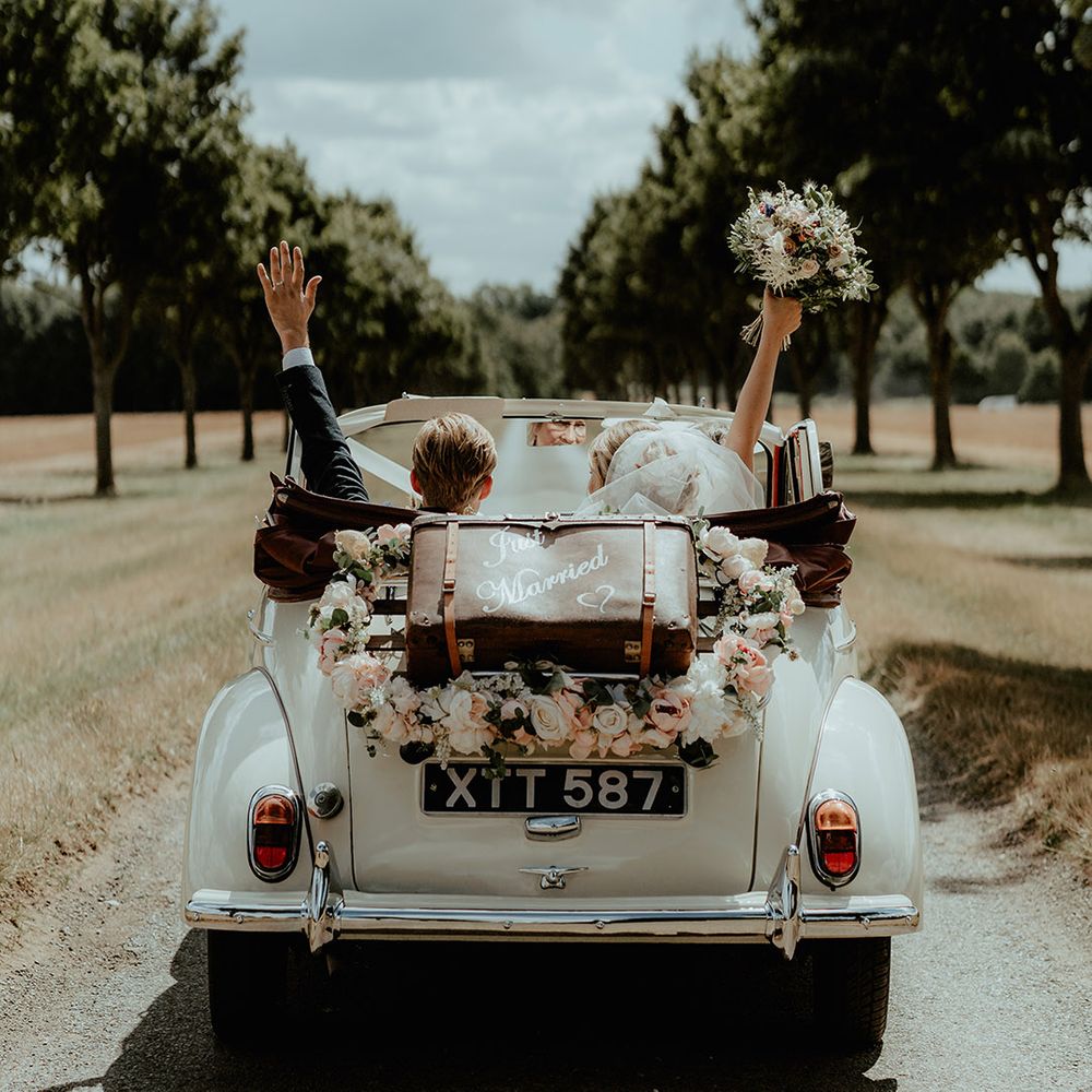 Couple ride away in a white vintage wedding car with an old vintage wedding suitcase with 'Just Married' written on it 