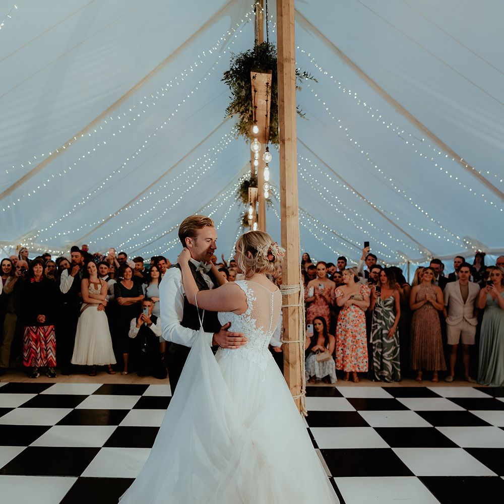 Bride and groom share their first dance with a checkerboard dance floor and fairy light decor and festoon lighting 
