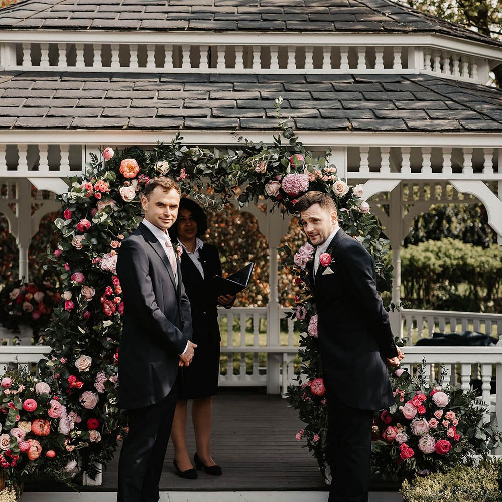Groom and best man stand waiting for the bride in front of the pink flower arch and other pink florals for the outdoor ceremony 