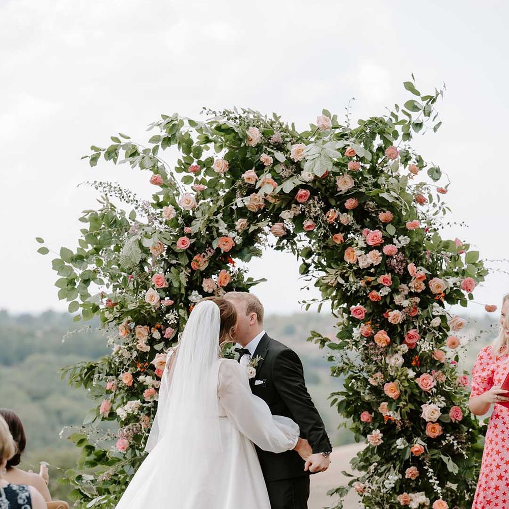 Bride wearing full sheer sleeve wedding dress kisses her groom in front of floral archway filled with orange and pink roses 