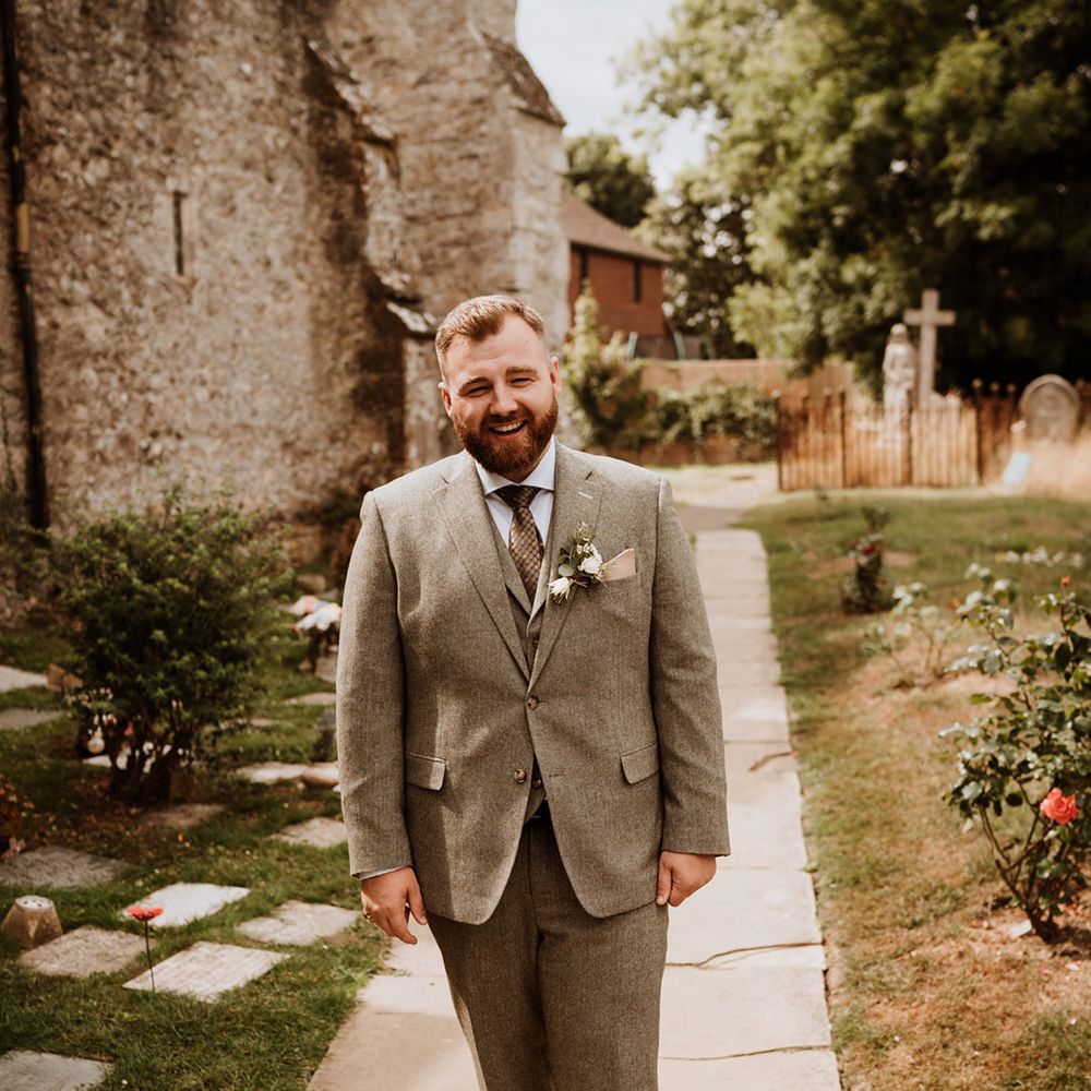 Bearded groom stands smiling in a three piece grey tweed suit 