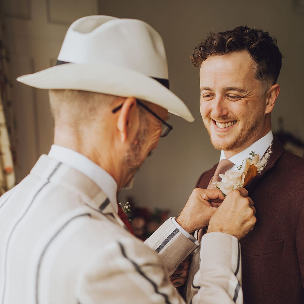 Groom has his buttonhole placed on maroon suit on the morning of his wedding day