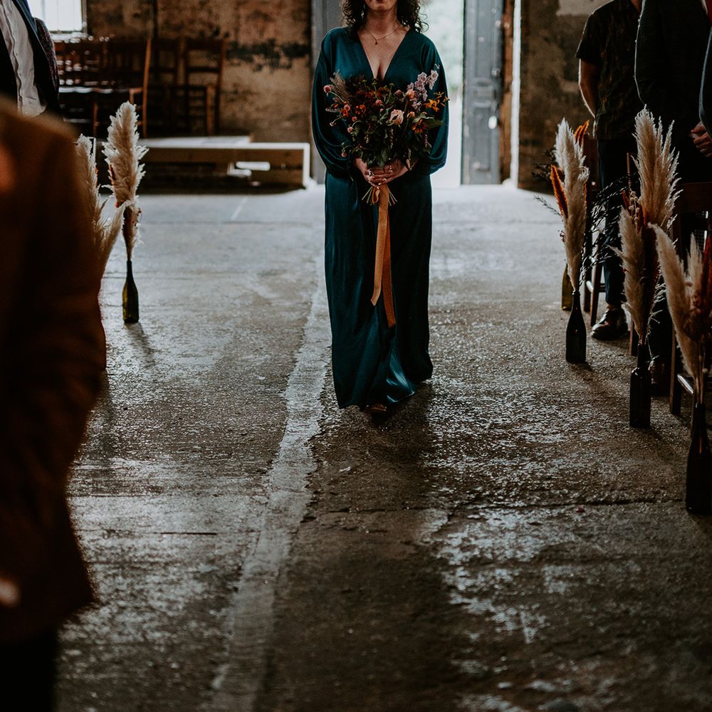 Bridesmaid walks down the aisle in green satin bridesmaid dress, carrying autumnal wedding flowers