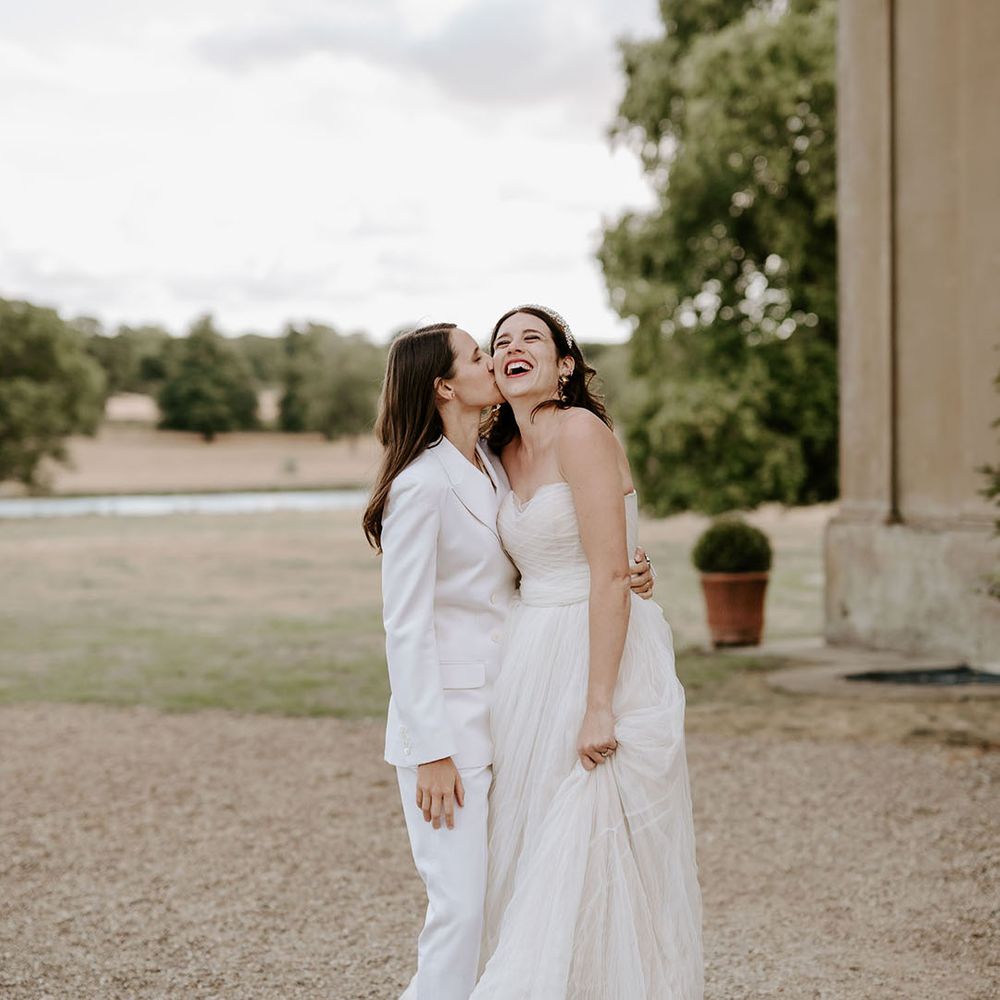 Bride in off the shoulder tulle wedding dress gets a kiss on the cheek from the bride in a white suit 