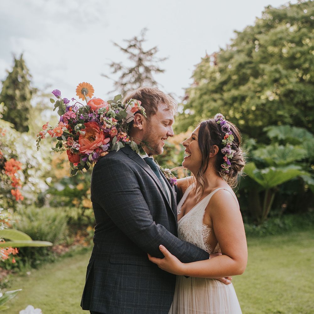 Bride wears colourful flower crown and scallop lace wedding dress whilst embracing her groom outdoors 