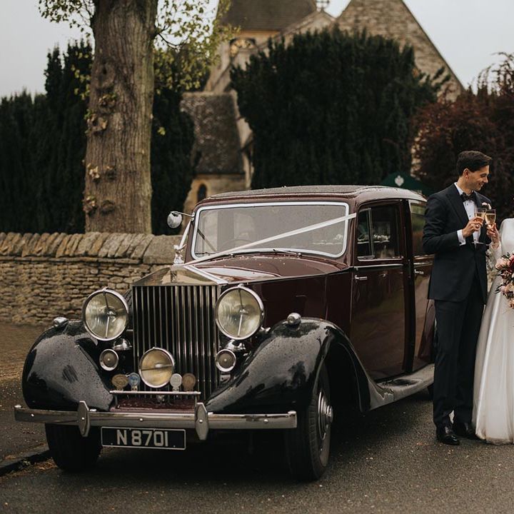 The bride and from drink champagne together and share a toast next to the vintage bridal car 