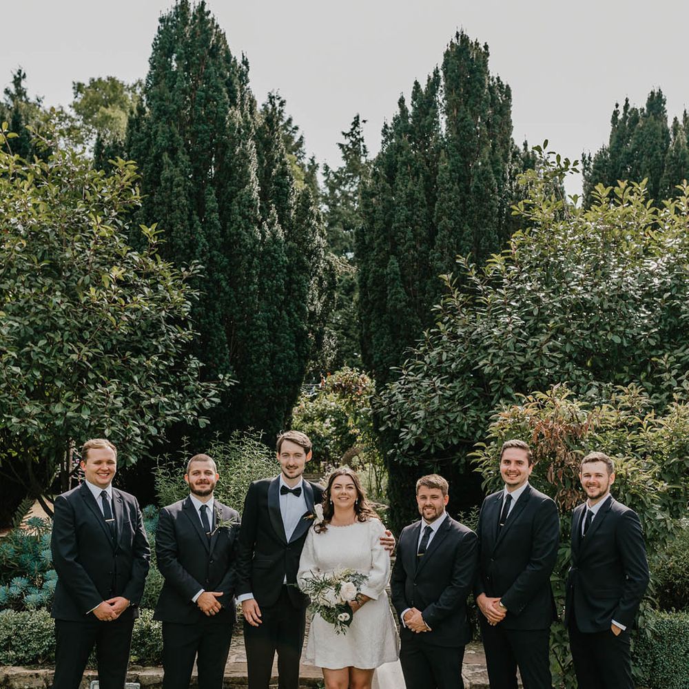 Groomsmen in black and white suits with the groom in a black tuxedo smiling with the bride in a short wedding dress with bow and train 