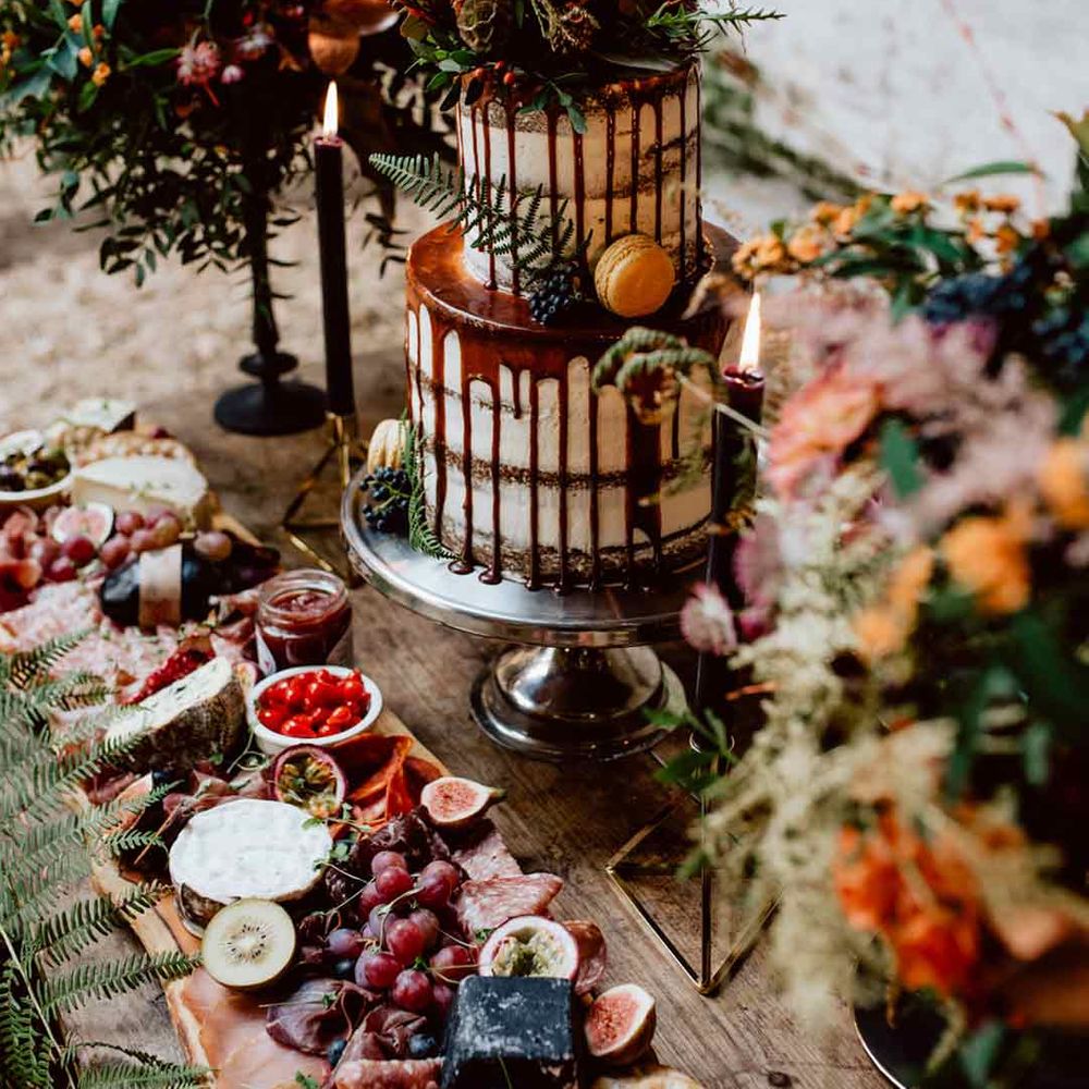 Two tier buttercream wedding cake with chocolate drip icing and dried flower decorations with dark tapered candles and a platter of fruit 