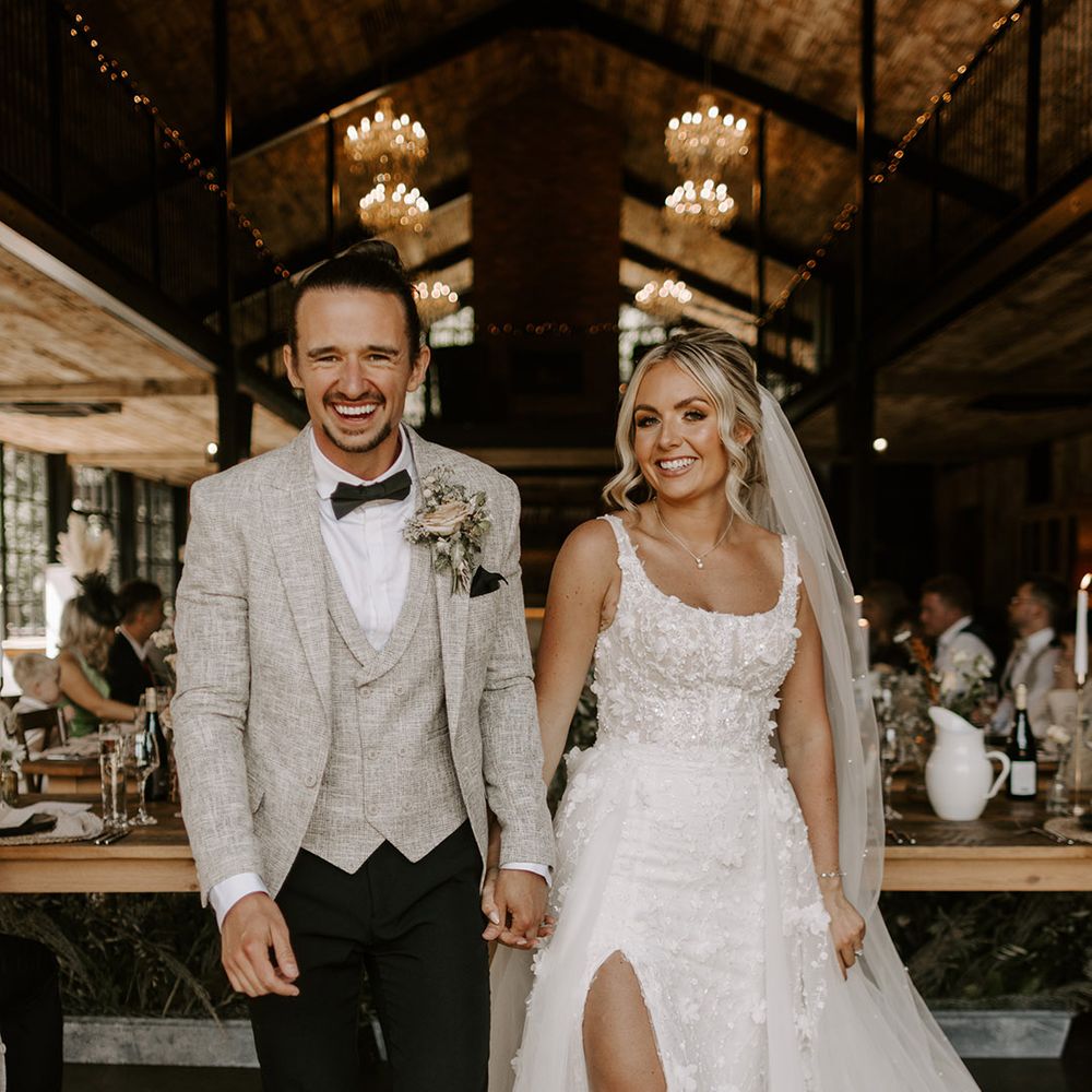 The bride in a split let embellished floral bridal gown walking with the groom in a light grey three piece suit with floral buttonhole at Hidden River Cabins