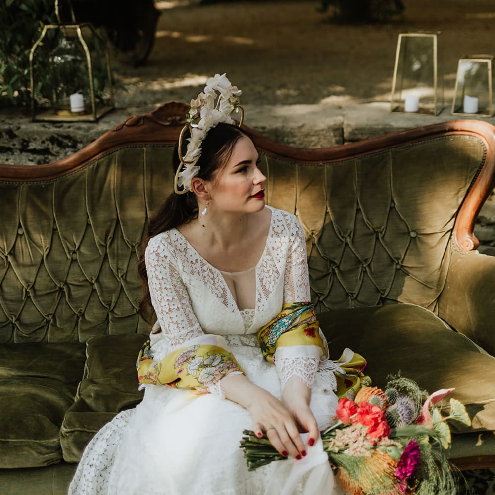 Bride in lace wedding dress and bridal crown with bridal bouquet consisting of red pincushion flowers, coral carnations, magenta peonies and anthuriums, sitting on a vintage olive green upholstered sofa