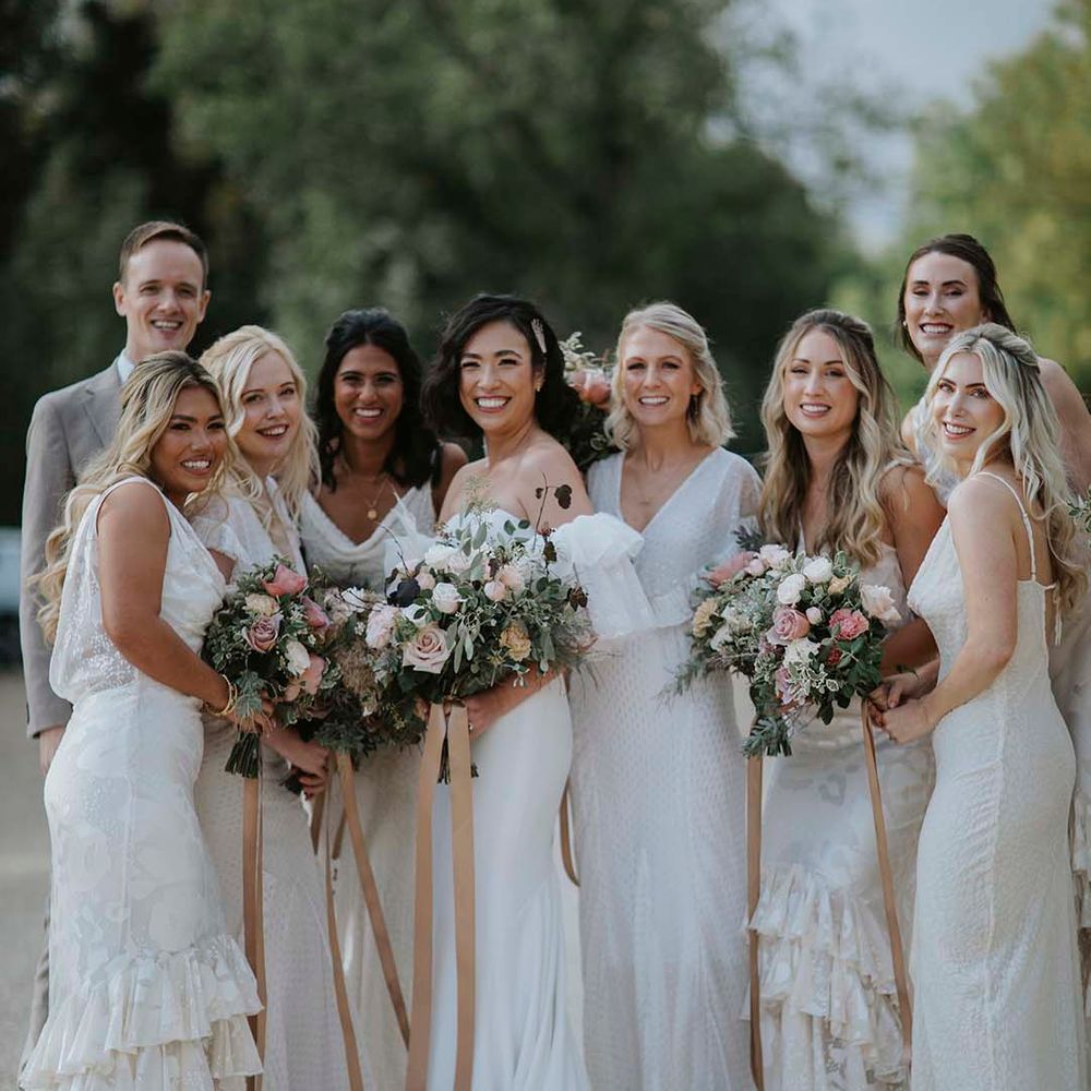 Bride in off the shoulder wedding dress standing with wedding party with bridesmaids in sleeveless layered white bridesmaid dresses 