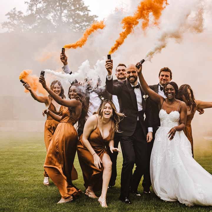 Bride in sweetheart neckline lace wedding dress with detachable off the shoulder sleeves, groom in classic black three piece grooms suit with bow tie and white rose and foliage boutonniere in church, bridesmaids wearing strappy sleeveless front slit burnt orange bridesmaid dresses holding foliage bouquets and groomsmen wearing classic black suits doing burnt orange and white smoke flares at Lains Barn wedding venue