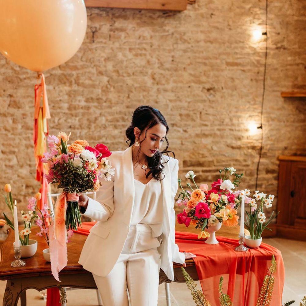 Bride in white bridal suit with closed toe bright green wedding heels standing by wooden table with orange table runner and mixed seasonal flower arrangements at Merriscourt wedding venue