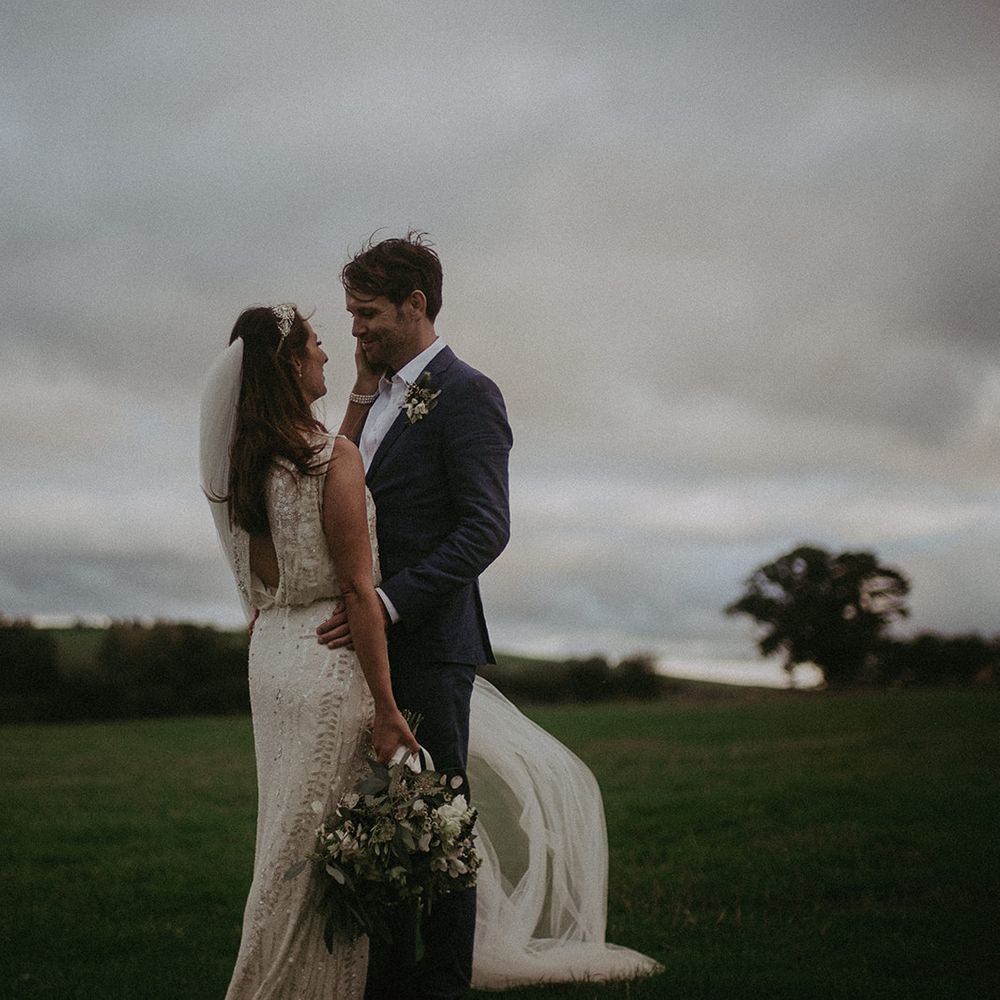 Bride & groom stand together outdoors in green fields on their wedding day