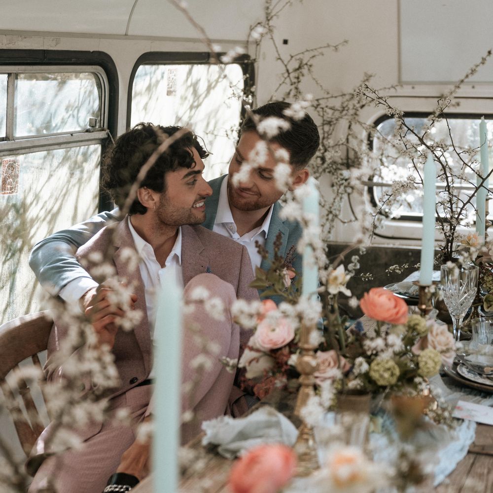 Grooms sit amongst rustic floral displays on their wedding day whilst looking lovingly at one another