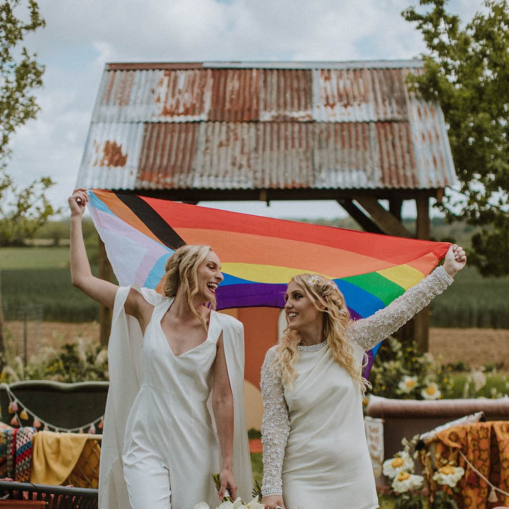 LGBTQI+ couple in a short wedding dress and bridal jumpsuit holding a colourful Pride flag 