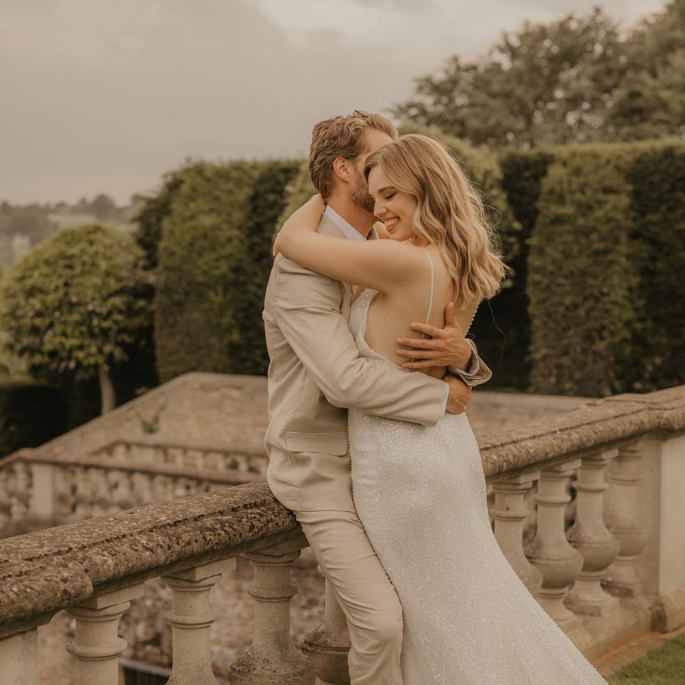 Groom in a beige suit and tan brogues embracing his bride in a sparkly Made With Love wedding dress on the terrace at Euridge Manor 