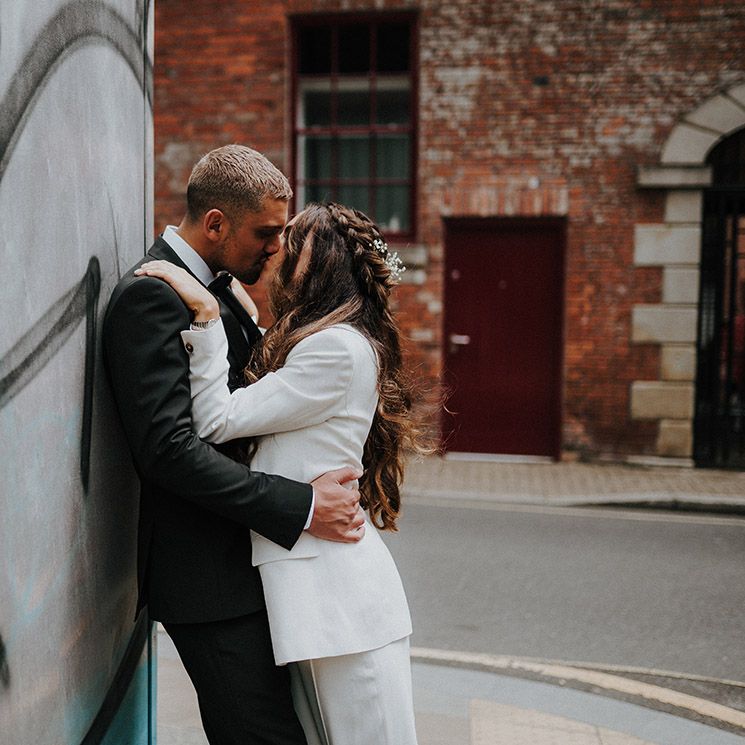 Bride with long wavy half up half down hairstyle in a white suit kissing her groom in a tuxedo 