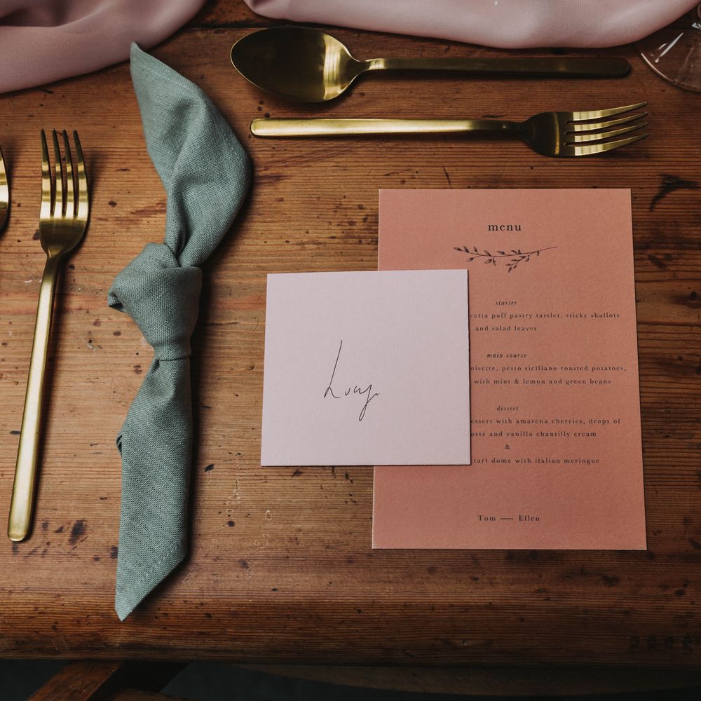 Pink & white stationery on wooden table next to sage green cloth napkin and gold cutlery