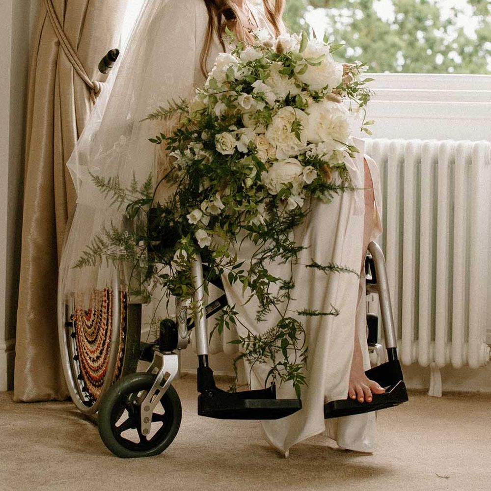 Wedding morning bridal preparations with bride in wheelchair wearing a getting ready robe holding her white and green trailing wedding bouquet 
