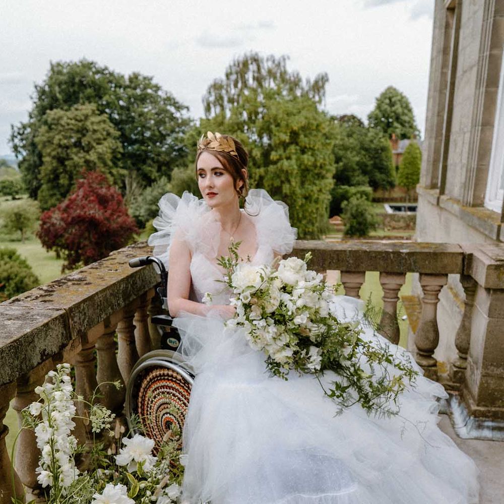 Portrait of the bride in a wheelchair in a tulle wedding dress with gold crown holding a white and green wedding bouquet on the balcony at Bourton Hall 