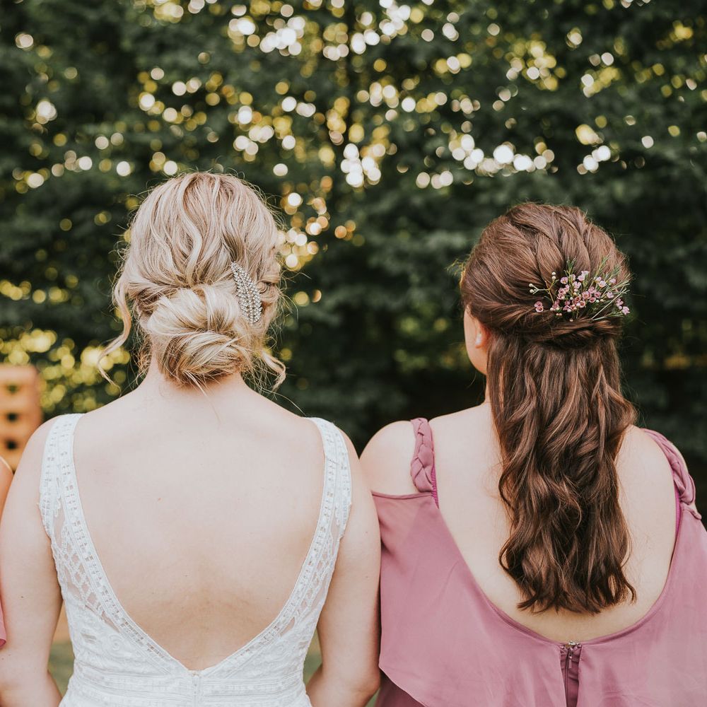 Bride with plaited bun in low v back Justin Alexander wedding dress stands in between bridesmaids with curled hair in dusky pink bridesmaid dresses outside at Tythe Barn wedding with barn wedding flowers