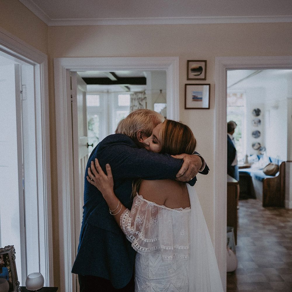 Bride in Grace Loves Lace bardot wedding dress and veil hugs man in blue suit in hallway before Isle of Wight wedding with macrame wedding decor