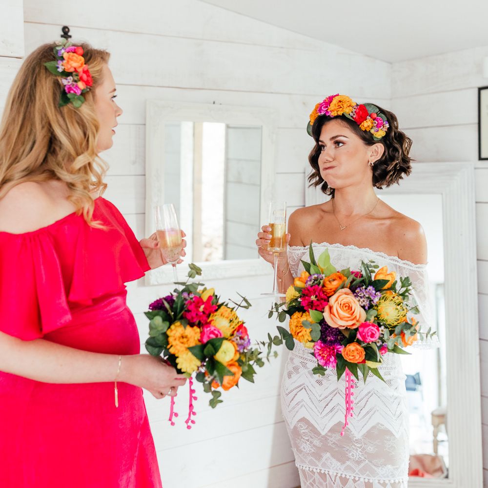 Bride in off the shoulder Grace Loves Lace wedding dress with colour flower crown and colourful bridal bouquet stands with bridesmaid in pink bridesmaid dress as hey both hold glasses of sparkling wine