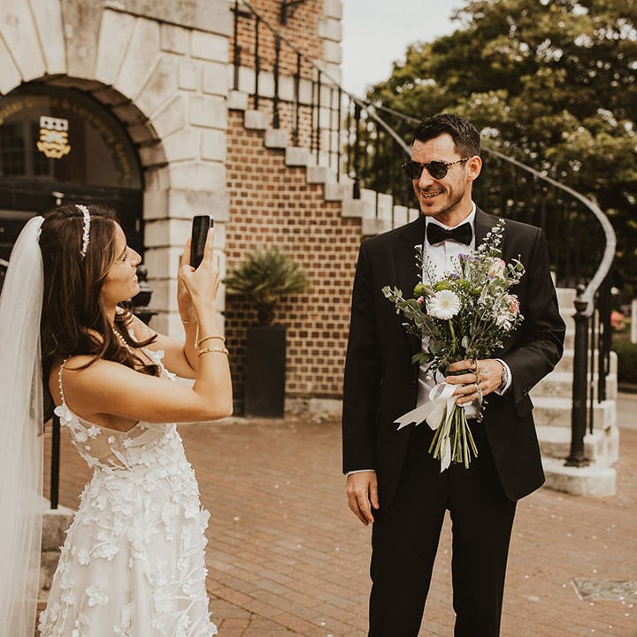 Bride takes photo of the groom who holds her floral bouquet and wears sunglasses 