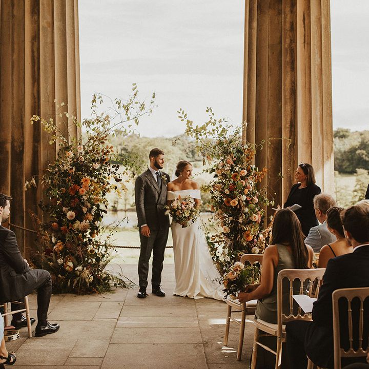 Bride & groom stand in front of floral archway next to large pillars outdoors during wedding ceremony