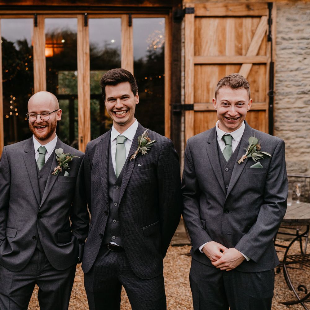 Groom and groomsmen in grey three piece wedding suits, green ties and thistle buttonholes stand outside barn after wedding ceremony