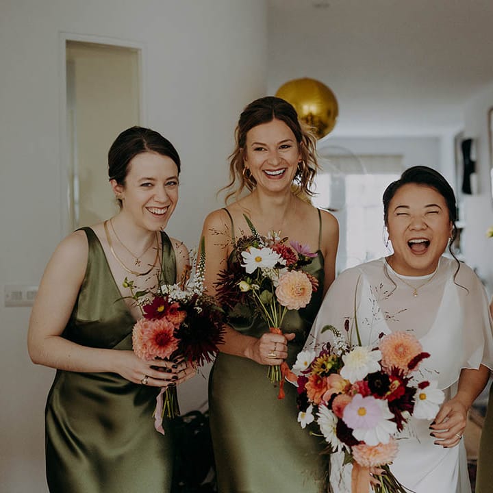 Bride laughs with her bridesmaids as they hold bright floral bouquets and bridesmaids wearing olive green satin dresses  | Irene Yap Photography