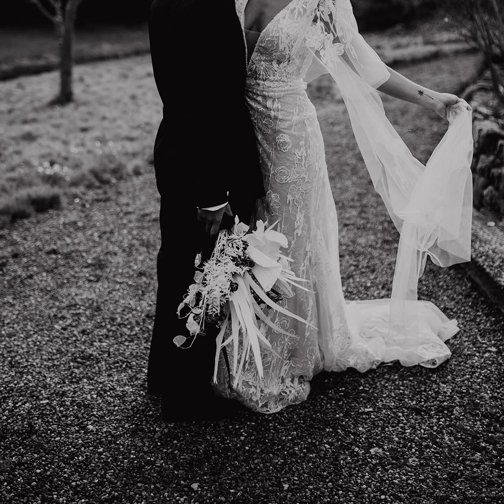 Bride in a lace wedding dress with lunging neckline and watteau train embracing her partner in a black suit 