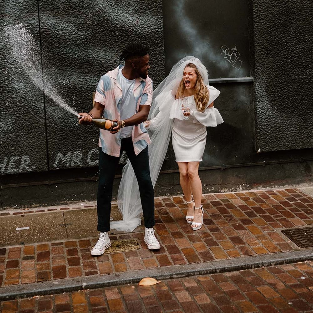 Groom in a pastel patterned shirt and Converse trainers popping champagne with his bride in a short wedding dress and long veil