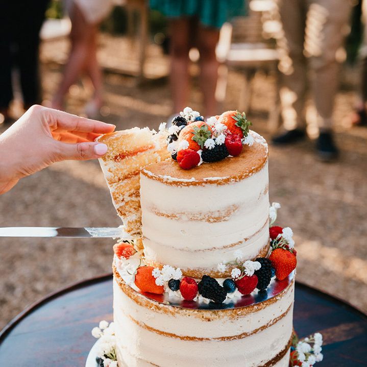 Bride takes a slice of cake from two tier semi naked white frosted wedding cake with summer berries and gypsophila 