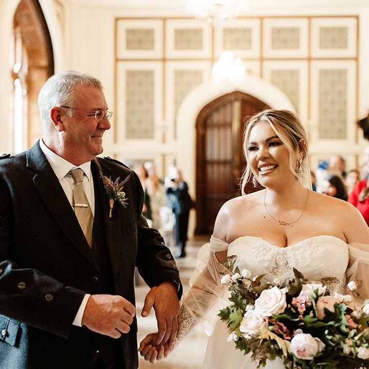 Father of the bride in dark suit jacket and beige tie with silver tie clip gives away the bride in strapless wedding gown 