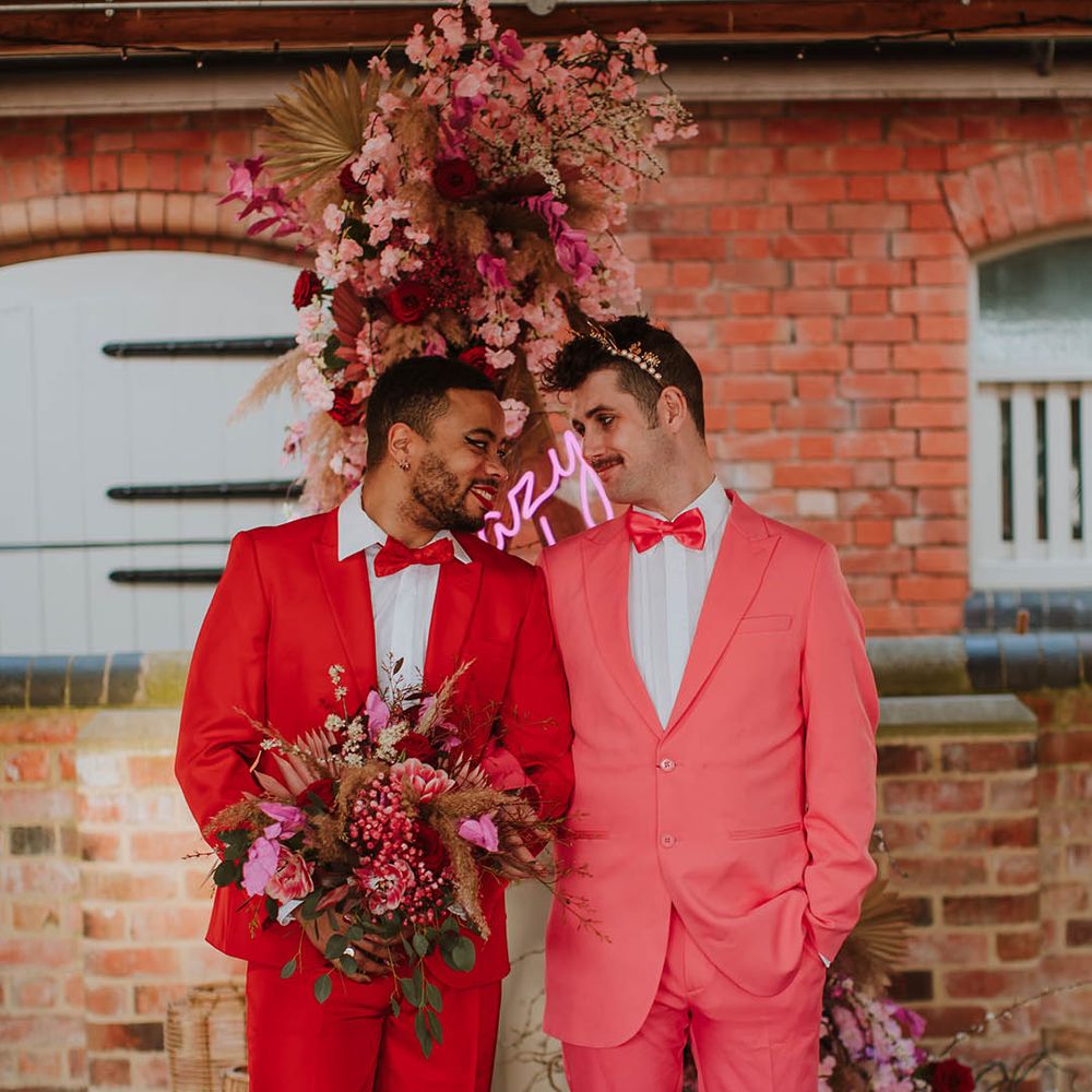 Groom in a red suit holding a pink bouquet with makeup on standing next to his partner in a pink suit, bow tie and gold crown