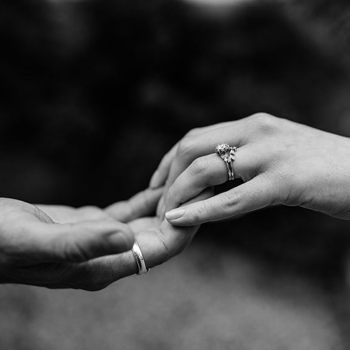 Bride and groom hold hands to show off their rings with bride wearing wishbone wedding ring and round engagement ring and groom's plain wedding band 