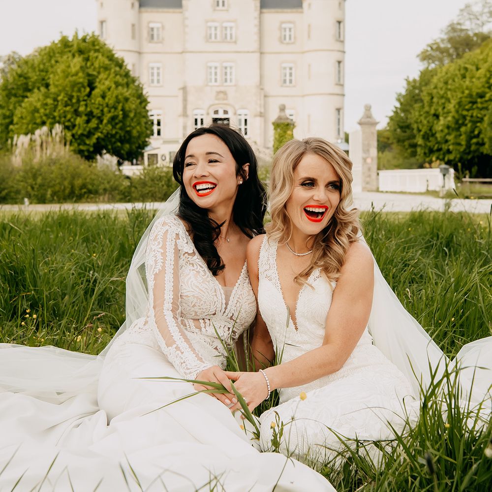 Brides laugh together as they sit in the grass outside their wedding venue 