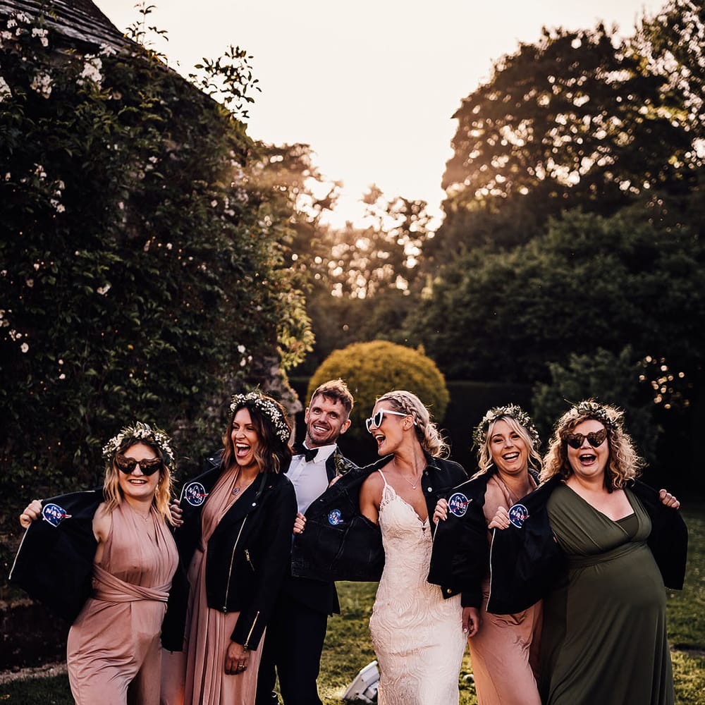 Bride in retro heart sunglasses with bridesmaids in pink and green dresses and groom all in matching NASA leather jackets 