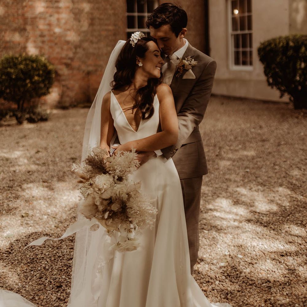 Bride in v neck wedding dress from Alexandra Grecco is embraced by the groom from behind 