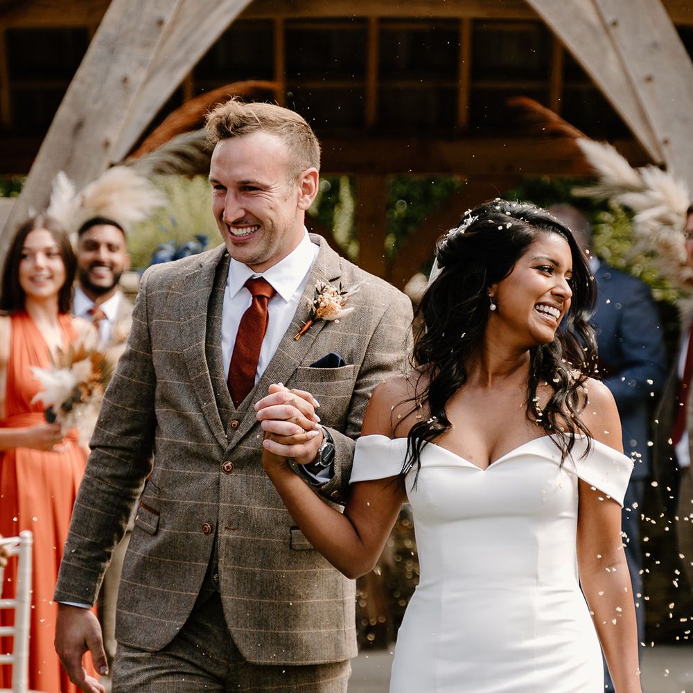 Bride in an off the shoulder fitted wedding dress walks through confetti with the groom in a three piece brown suit and orange tie 