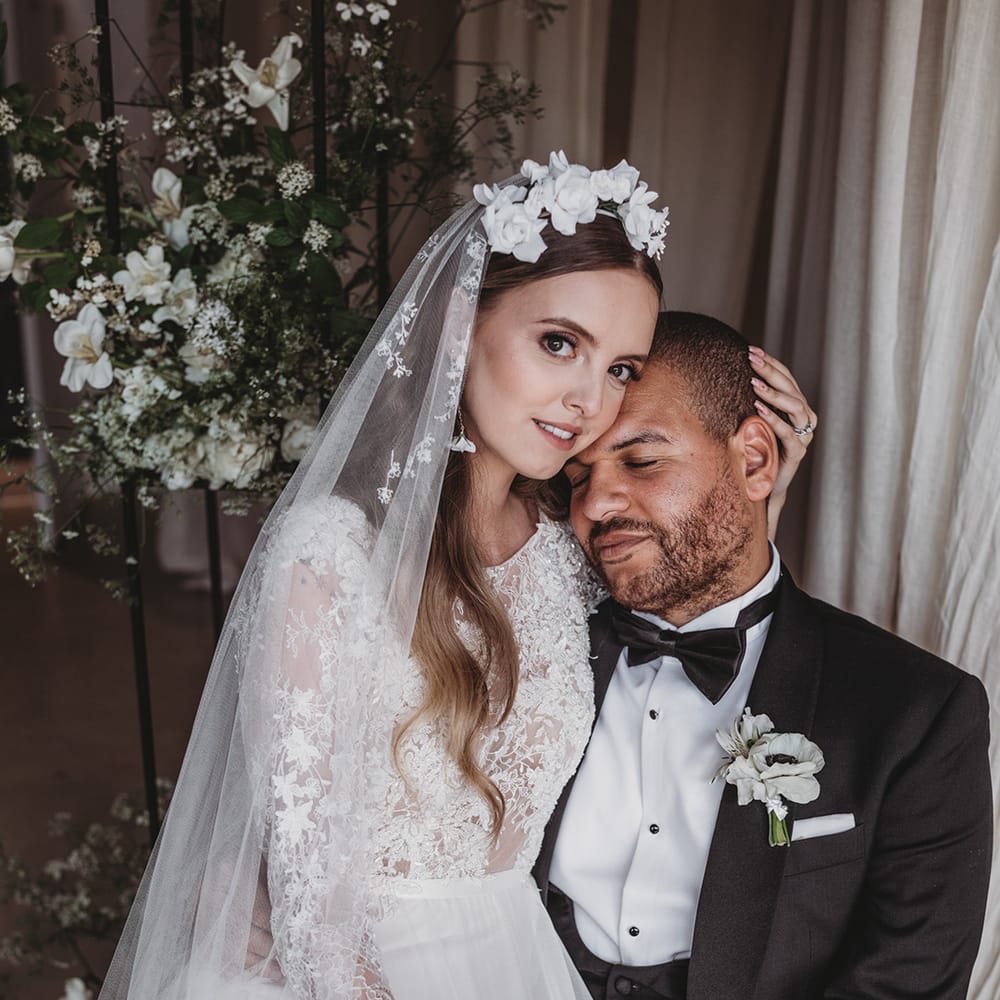Bride with an embroidered veil, flower headband and earrings in a lace wedding dress cups the groom's head towards her