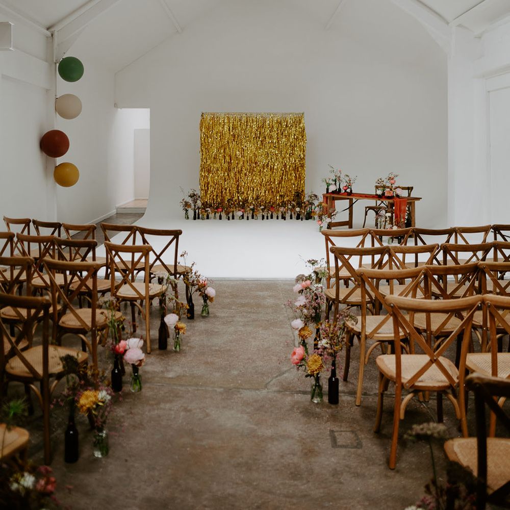 Gold streamers wedding decor for industrial styled wedding with wooden chairs and small floral arrangements lining the aisle