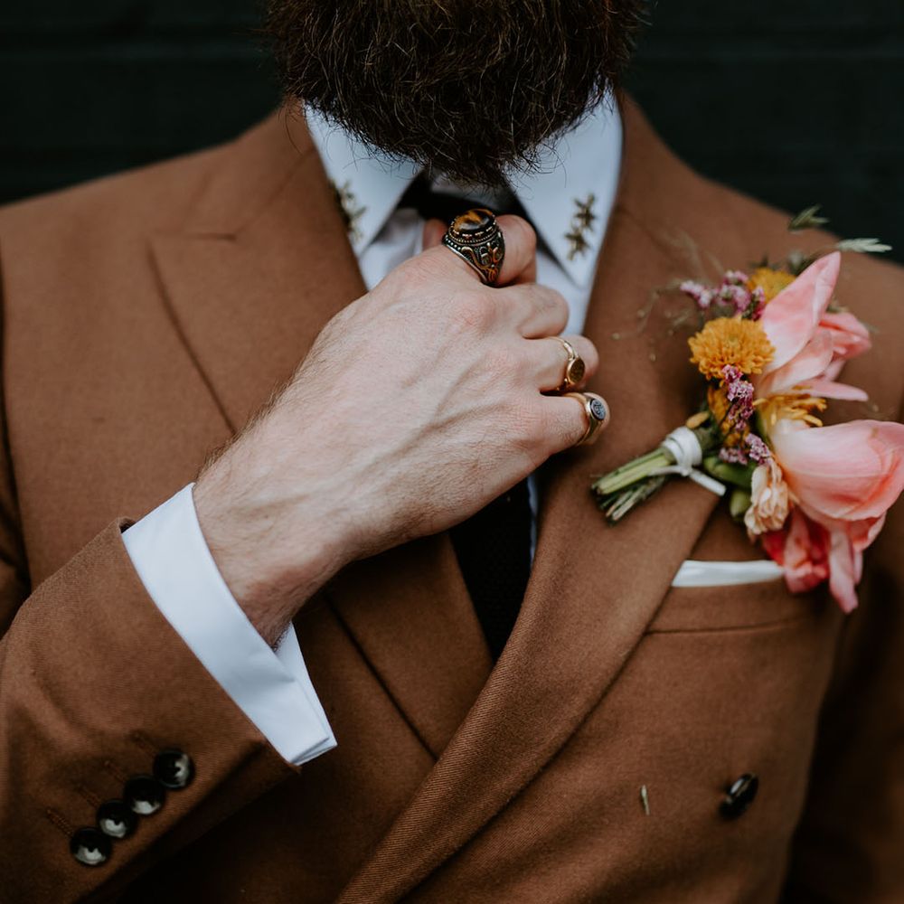 Groom wears brown retro suit complete with pink floral buttonhole and wears silver statement rings
