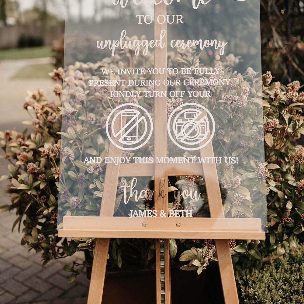 Clear acrylic wedding welcome sign on a wooden easel with pink flowers and foliage 